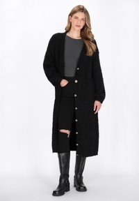 Junge Frau mit langen Haaren, die einen schwarzen, kuscheligen langen Cardigan, ein graues Top, zerrissene schwarze Jeans und schwarze kniehohe Stiefel trägt und vor einem weißen Hintergrund steht.