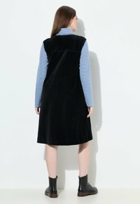 Schwarzes Cordkleid mit ärmellosem Design, Rückennahtdetails und weitem Rock, kombiniert mit einem blau-weiß gestreiften Langarmtop und Ankle Boots.