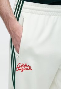 Άσπρο παντελόνι φόρμας με πράσινες ρίγες στα πλευρά, διακοσμημένο με κεντημ ένο κόκκινο λογότυπο «adidas Originals» και πλαϊνή τσέπη με φερμουάρ.