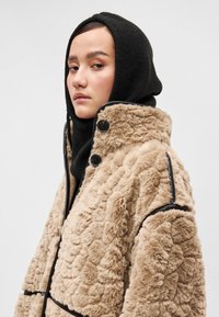 Bežs mākslīgā kažokādas jaka ar teksturētu virsmu, ar melniem akcentiem un pogām. Valkāta virs melnas adītas šalles.