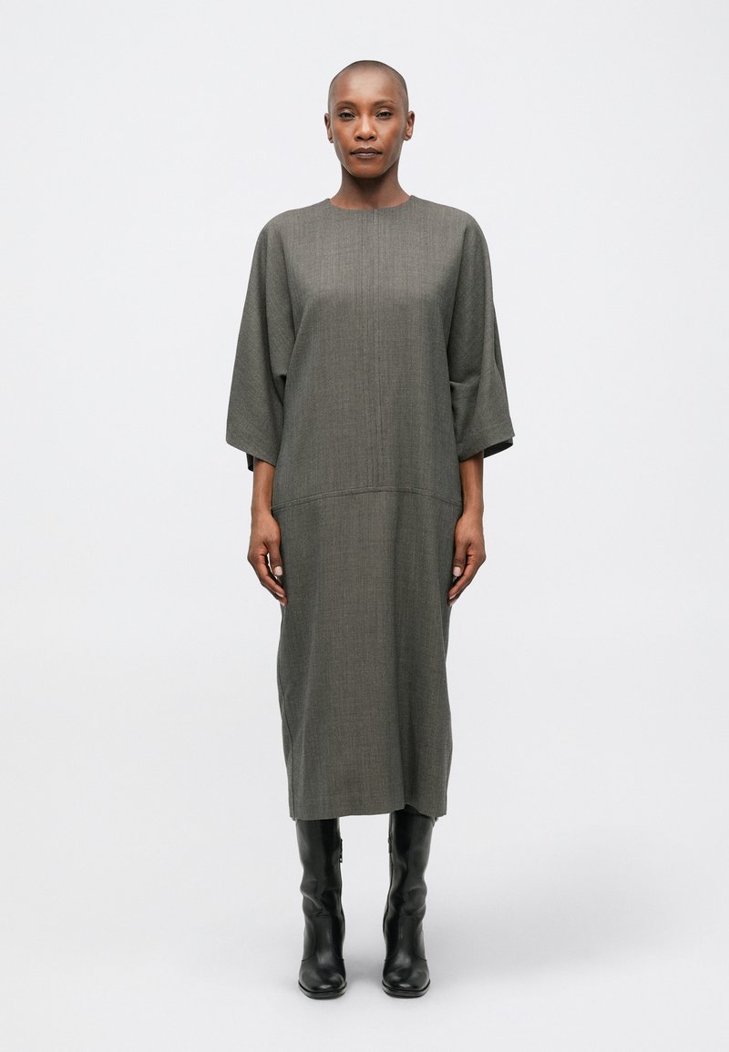 Robe grise, mi-longue, avec une coupe droite et des manches larges ; présente un tissu texturé et un col rond. Portée avec des bottes noires jusqu'aux genoux.