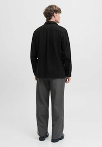 Camicia nera con bottoni realizzata in tessuto testurizzato, caratterizzata da un colletto tradizionale, maniche lunghe e un taglio dritto. Indossata con pantaloni grigi.