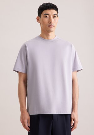 Młody mężczyzna z krótkimi czarnymi włosami, ubrany w gładki, jasnofioletowy t-shirt z krótkim rękawem i ciemne spodnie, stoi na tle o neutralnym kolorze.