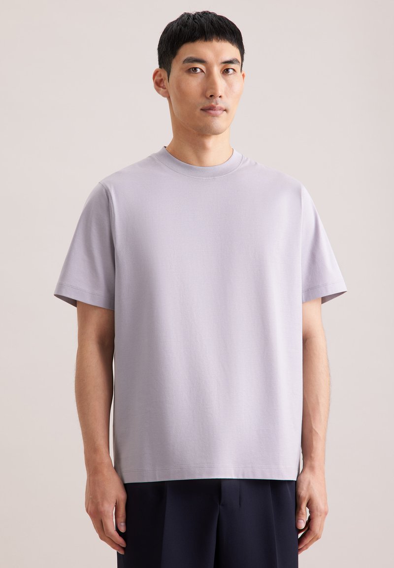 Jeune homme aux cheveux noirs courts portant un t-shirt uni à manches courtes couleur lavande claire et un pantalon foncé, debout devant un fond neutre.