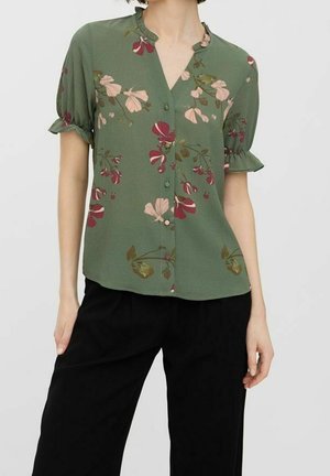 Blouse verte à manches courtes avec imprimé floral rose et rouge, boutonnée sur le devant, poignets à volants, portée avec un pantalon noir ample sur une personne debout.