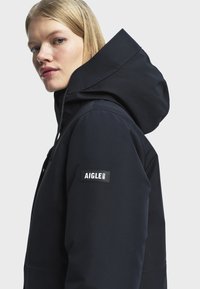 Femme blonde portant une veste à capuche noire avec un patch logo "AIGLE 1853" visible sur la manche gauche, regardant légèrement par-dessus son épaule.