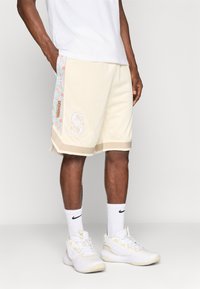 Beige sporta šorti ar sieta audumu, gaiši zilo rakstoto sānu paneļu ar ziedu dizainu, ar logotipu un kontrastējošu apdari.