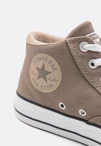 Brązowy sportowy but w stylu high-top z białymi sznurówkami i czarną gumową podeszwą. Posiada okrągłą beżową naszywkę z logo z gwiazdką, oznaczoną "Converse All Star."