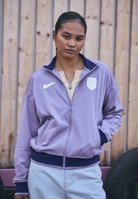 Lilla zip-up jakke med marineblå detaljer, Nike-logo og et skjoldpatch. Mykt stoff og elastisk liv, med sidelommer og ståkrage.