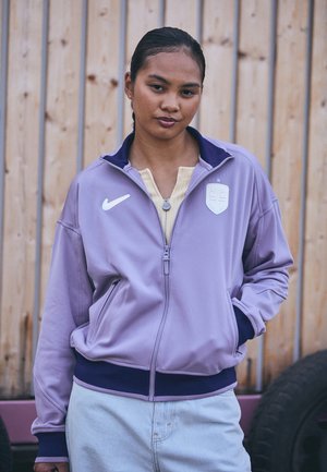 Veste zippée violette avec des accents marine, logo Nike et écusson. Tissu doux et taille élastique, avec des poches latérales et un col montant.