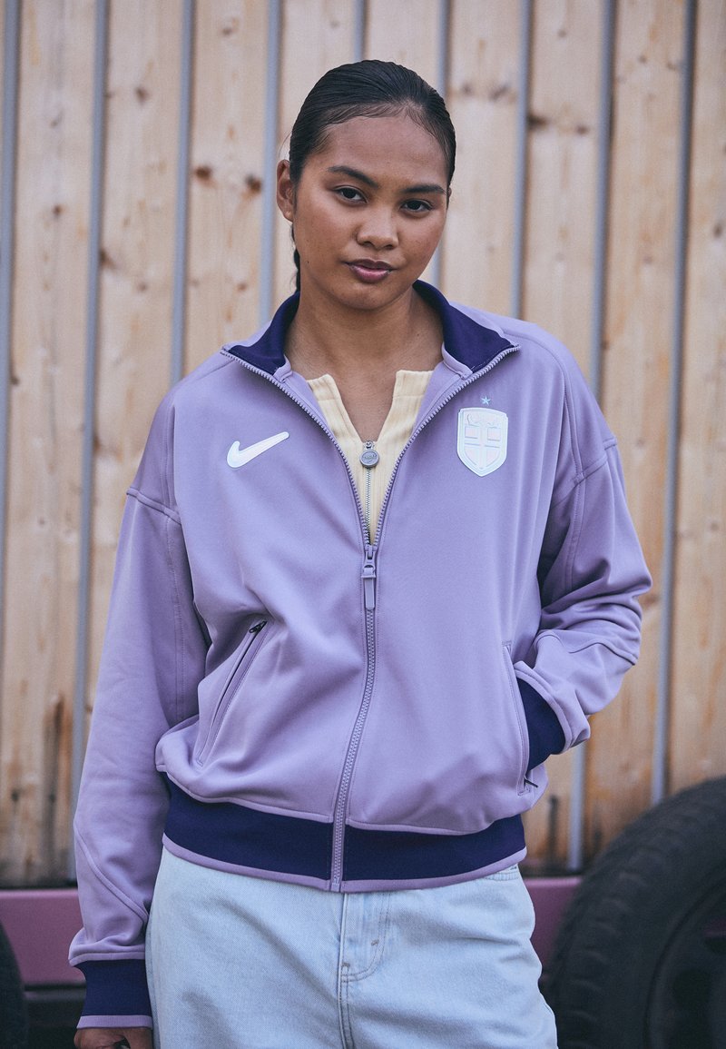 Lilla zip-up jakke med marineblå detaljer, Nike-logo og et skjoldpatch. Mykt stoff og elastisk liv, med sidelommer og ståkrage.