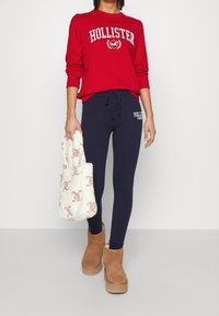 Sudadera roja con el logo de "Hollister", leggings navy, botas de tobillo beige y un bolso blanco esponjoso con un patrón repetido. Look casual.