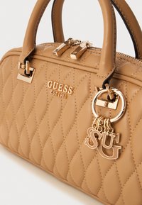 Geantă din material quiltat de culoare bej, cu accesorii aurii, închidere cu fermoar și un charm decorativ cu literele "S" și "G." Suprafață din piele texturată.