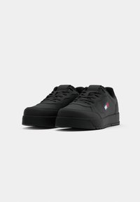 Paire de baskets basses noires avec lacets, semelles épaisses et un petit logo rectangulaire rouge, blanc et bleu sur le côté.