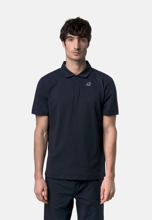 Uomo con capelli scuri che indossa una maglietta polo nera a maniche corte con un piccolo logo bianco sul petto a sinistra, su uno sfondo semplice.