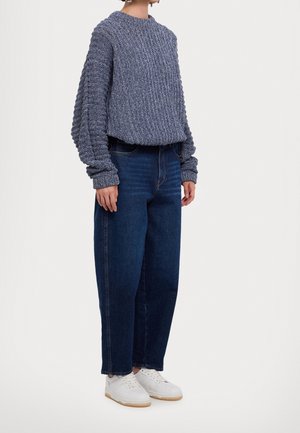 Person iført en kraftig blå striktrøje, mørkeblå højhægtede jeans og hvide sneakers, stående mod en ensfarvet baggrund.