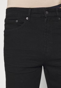 Jeans in denim nero con vestibilità slim. Le caratteristiche includono un'apertura con bottone, tasche frontali con rivetti metallici e una texture liscia.