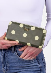 Green wallet with large yellow polka dots, gold hardware, and a zipped closure. Smooth texture and rectangular shape, held in a hand.