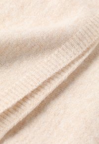 Beige gebreide stof met een zachte textuur, gekenmerkt door subtiele kleurvariaties en een ribbelrand langs de rand.