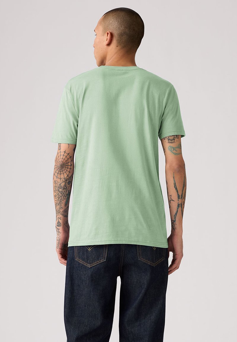 Mann mit kurzen Haaren und tätowierten Armen, der ein hellgrünes kurzärmeliges T-Shirt und dunkelblaue Jeans trägt, blickt von hinten.