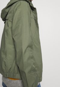 Olivengrøn letvægtsjakke med lynlås, hætte og elastiske manchetter. Har en glat tekstur og sidelommer.