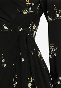 Svart klänning med blommönster som har vita och gula blommor. Den har en knutdetalj vid midjan och draperad tyg.