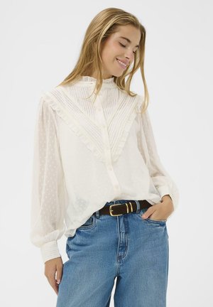 Hvid, semi-gennemsigtig bluse med flæsekrave, lodrette striber og prikker. Lange ærmer med knapmanchetter; parret med højtaljede denim jeans.