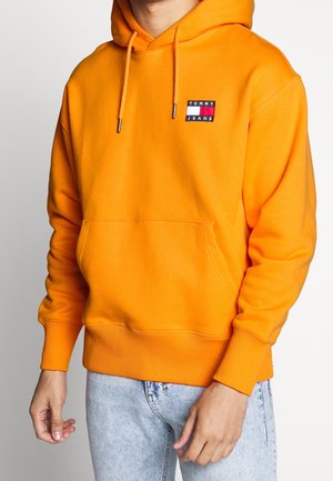 Man draagt een feloranje Tommy Jeans hoodie met voorzak en lichtblauwe jeans, staand tegen een effen achtergrond.