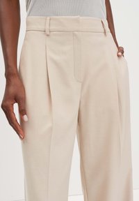 Beige op maat gemaakte broek met een geplisseerde voorkant, zijzakken en riemlussen. Gemaakt van een gladde stof met een lichte glans.