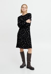 Robe noire à fleurs avec des manches longues, présentant une coupe relaxed et un motif de petites fleurs violettes et vertes. Accompagnée de bottes marron hautes.