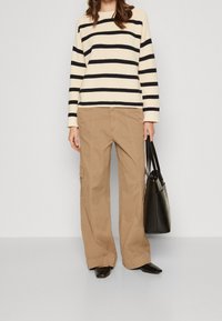 Maglione a righe in crema e blu navy, abbinato a pantaloni cargo larghi color beige. Modella tiene una borsa a tracolla nera, indossando scarpe con tacco nere.