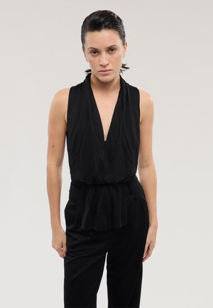 Donna con capelli tirati all'indietro che indossa un top nero senza maniche con scollo a V profondo e pantaloni neri, in piedi davanti a uno sfondo chiaro e semplice.