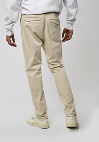 Pantaloni in cotone beige con una finitura testurizzata, dotati di due tasche posteriori e un design slim fit. Abbinati a sneakers bianche.