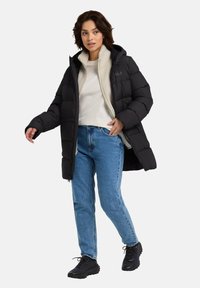 Piumino nero con cappuccio, dotato di una fodera in pile crema a contrasto, chiusura con zip e motivo trapuntato. Indossato sopra una camicia leggera e jeans.