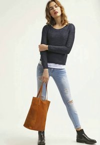 Navy gestrickter Pullover, abgewetzte hellblaue Jeans, schwarze Stiefeletten und eine orangebraune Tragetasche mit glatter Textur und klaren Linien.