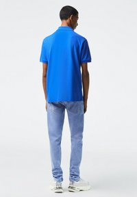 Blaues Polohemd, kurze Ärmel, aus Baumwolle. Kombiniert mit hellblauen Jeans und weißen Sneakers, von hinten betrachtet.