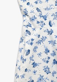 Hvidt stof med et blåt blomsterdesign i varierende nuancer. Designet inkluderer let struktur og dekorative udsparinger langs sømme.
