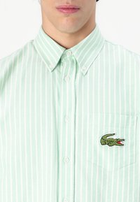 Light green striped button-up shirt featuring a pocket with an embroidered crocodile logo. Made from fabric with a smooth texture.
