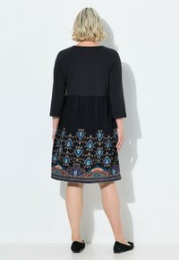 Schwarzes knielanges Kleid mit Dreiviertelärmel, das farbenfrohe geometrische Stickerei am Saum und einen lockeren Schnitt aufweist. Weiche Stofftextur.