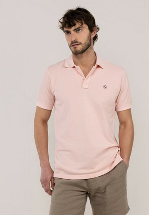 Hombre con cabello oscuro que lleva una camiseta polo rosa claro y pantalones cortos beige, de pie frente a un fondo liso.