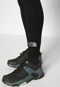 Leggings sportivi neri con pannelli laterali testurizzati, abbinati a scarpe da trail di colore nero e turchese con suola in gomma resistente e sistema di lacci.