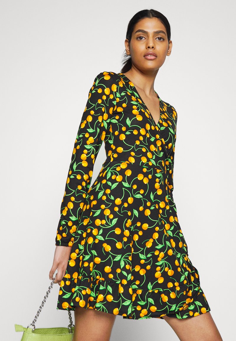 Vestido preto de mangas longas, com um padrão vibrante de cerejas amarelas e folhas verdes. Barra rodada com detalhe de volantes, combinado com uma bolsa verde.