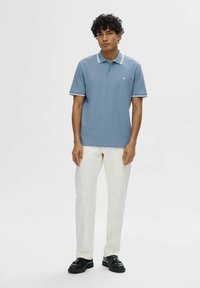 Polo shirt azzurro chiaro con rifiniture bianche a contrasto, maniche corte e un logo discret. Abbinato a pantaloni bianchi e scarpe nere.