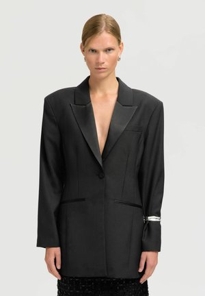 Femme aux cheveux slickés, portant une veste noire surdimensionnée avec des revers en satin et un bracelet en argent au poignet gauche, sur un fond clair.