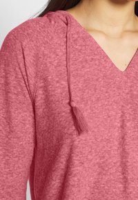 Roze pullover top met een V-hals ontwerp, gemaakt van zachte, gestructureerde stof. Heeft een capuchon met een franje detail aan het trekkoord.