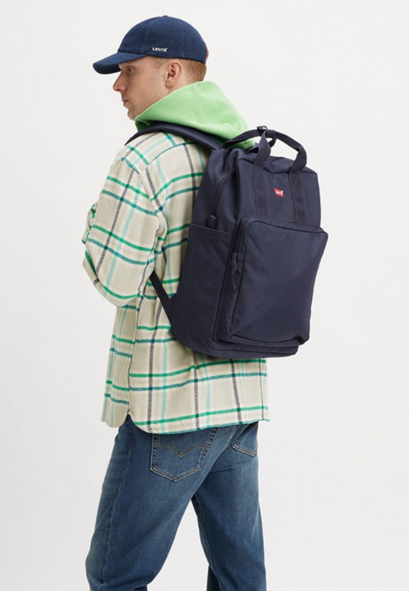 Navy mėlynas kuprinė su keliomis skyreliais ir viršutinėmis rankenomis, dėvima ant žalsvo gobtuvo ir languotos flanelinės marškinės. Patvari medžiaga.