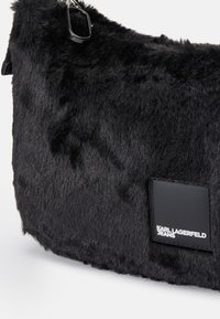 Μαύρη τσάντα ώμου από συνθετική γούνα με υφασμάτινη επιφάνεια, περιλαμβάνοντας γυαλιστερή μαύρη ετικέτα με λογότυπο. Διαθέτει μαύρο μεταλλικό εξάρτημα για τον ιμάντα.