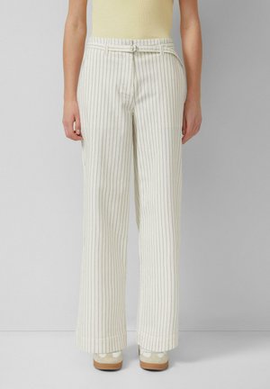 Persoon draagt witte wijde broek met dunne zwarte verticale strepen en een dunne witte riem, gecombineerd met witte sneakers.