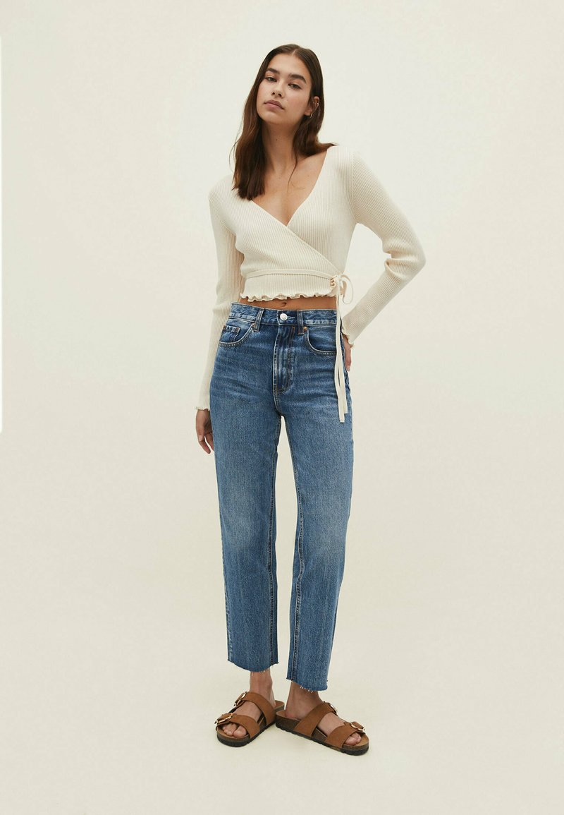 Top envolvente de canalé crema con escote en V y detalle de lazo, combinado con jeans de tiro alto en azul, cortos por encima del tobillo, y sandalias marrones.