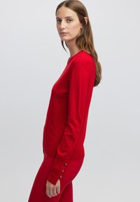 Maglione rosso a maniche lunghe con scollatura rotonda e dettagli di bottoni sui polsini, realizzato in un materiale morbido, dal design aderente.